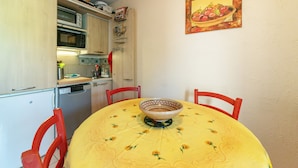 Een kleine keuken met een magnetron, oven en vaatwasser. Een eettafel met een tafelkleed met bloemenprint en een kom bevindt zich in het midden.