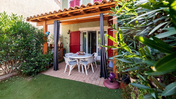 Een terras met een tafel en stoelen, omringd door groen en een gebouw met rode luiken.