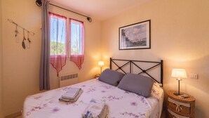 Een slaapkamer met een bed, twee bedlampjes, een raam met gordijnen, een wanddecoratie en een nachtkastje met een lamp.