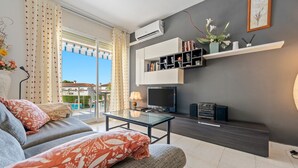 Un salón moderno con sofá, una mesa de centro de vidrio, un televisor de pantalla plana y un estante de pared con libros y objetos decorativos.