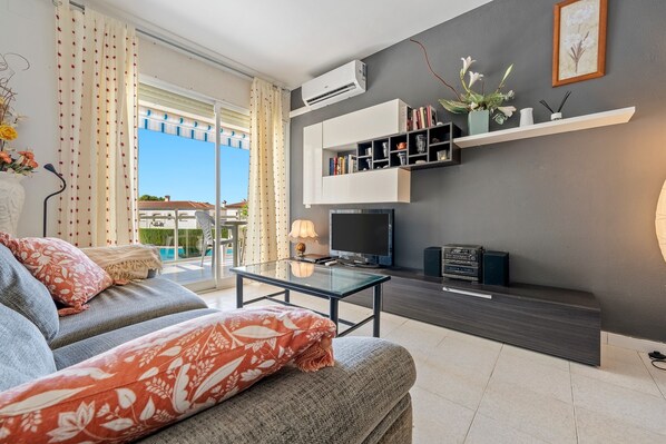 A modern living room with a sofa, a glass coffee table, a flat-screen TV, and a wall-mounted shelf with books and decorative items.