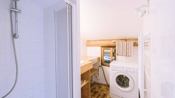 Une petite salle de bain comprenant une douche, un lavabo, un lave-linge et une étagère en bois.