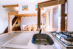 Una cocina con un lavaplatos de acero inoxidable, estufa a gas, vigas de madera, una escalera y un comedor con mobiliario de madera.