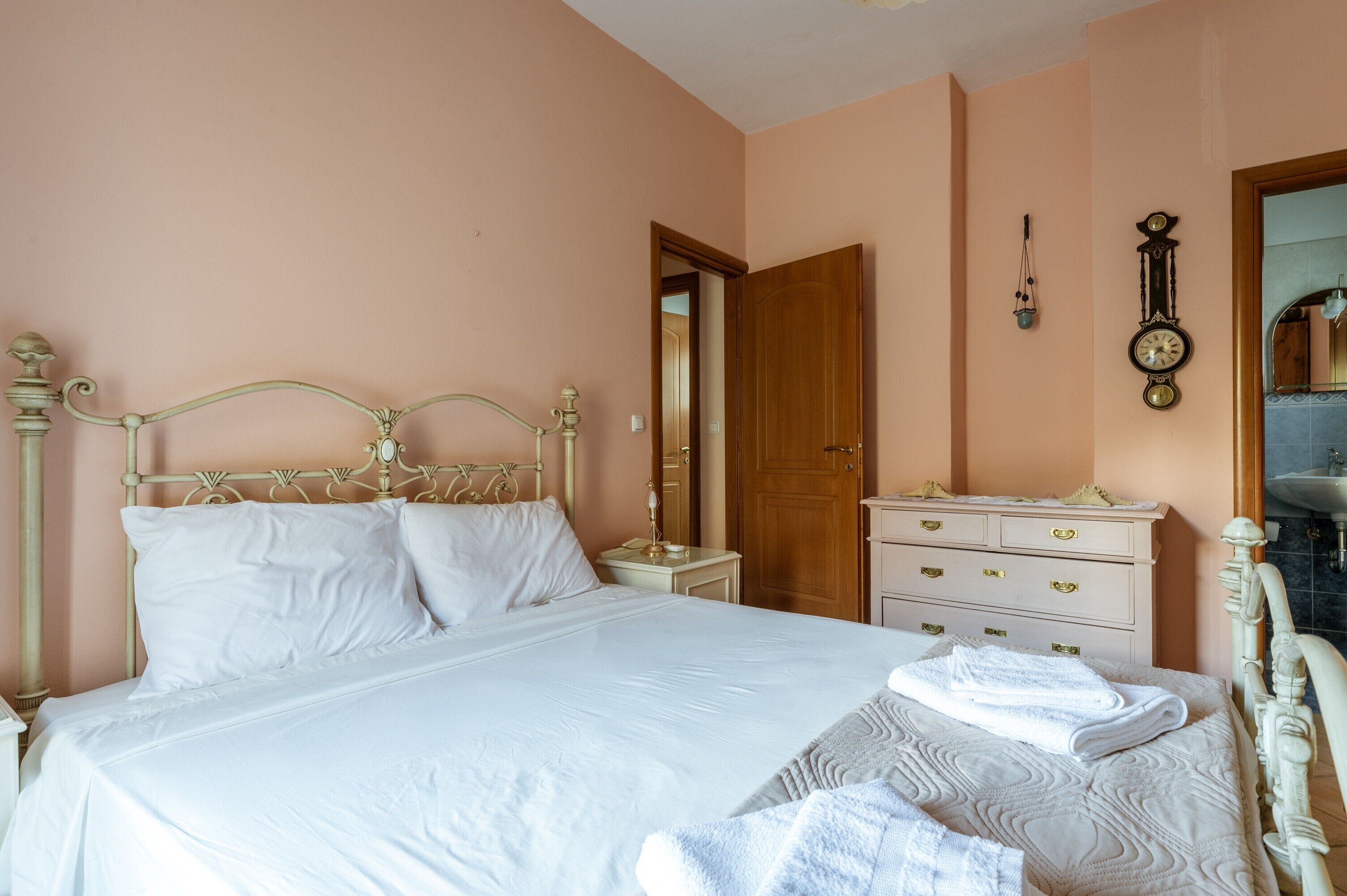 Una cama bien hecha con ropa de cama blanca, un aparador de madera con cajones, un reloj de pared y un espejo.