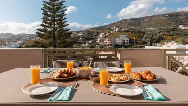 Une table dressée avec des aliments pour le petit-déjeuner, notamment des croissants et du jus d’orange, donnant sur une colline parsemée de bâtiments.