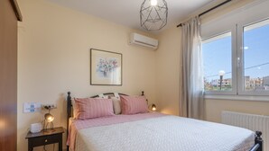 A bedroom with a bed, bedside table, lamp, air conditioning unit, window, and a city view. - Holiday Home 'Villa Melenia 1' with Private Pool, 5 Minutes from the Beach, Wi-Fi & Air Conditioning (??. ?e?a??a)