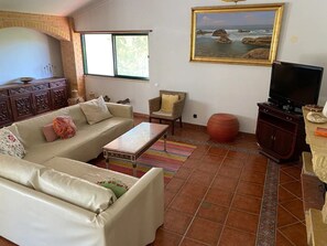 A living room with a sofa, armchair, TV stand, and a painting of a coastal scene.