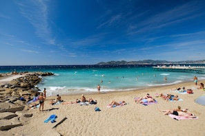 Una playa con gente tomando el sol y nadando, un muelle que se adentra en el agua, y un cielo azul despejado.