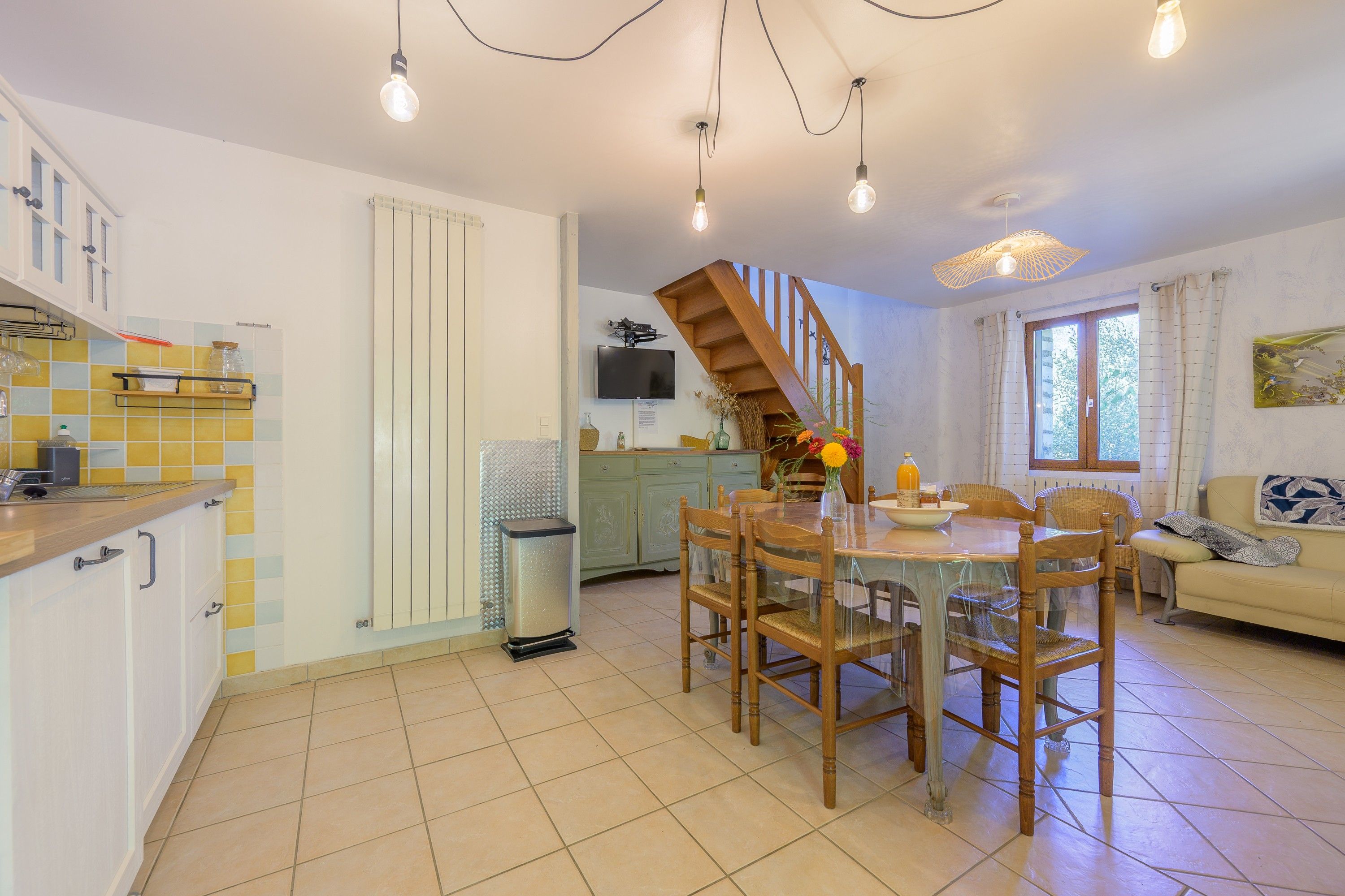 A spacious kitchen with a dining area, a staircase, and a living room with a sofa and a window.