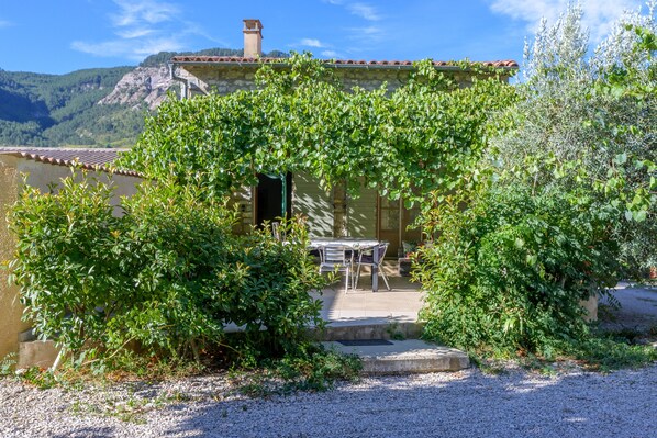 A rustic building with a stone facade, a tiled roof, and a gravel pathway leading to a table and chairs set up outside.