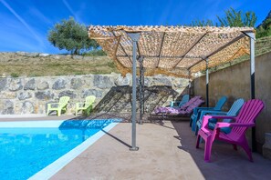 A pool area with a stone wall, a pergola with a patterned roof, and colorful lounge chairs.