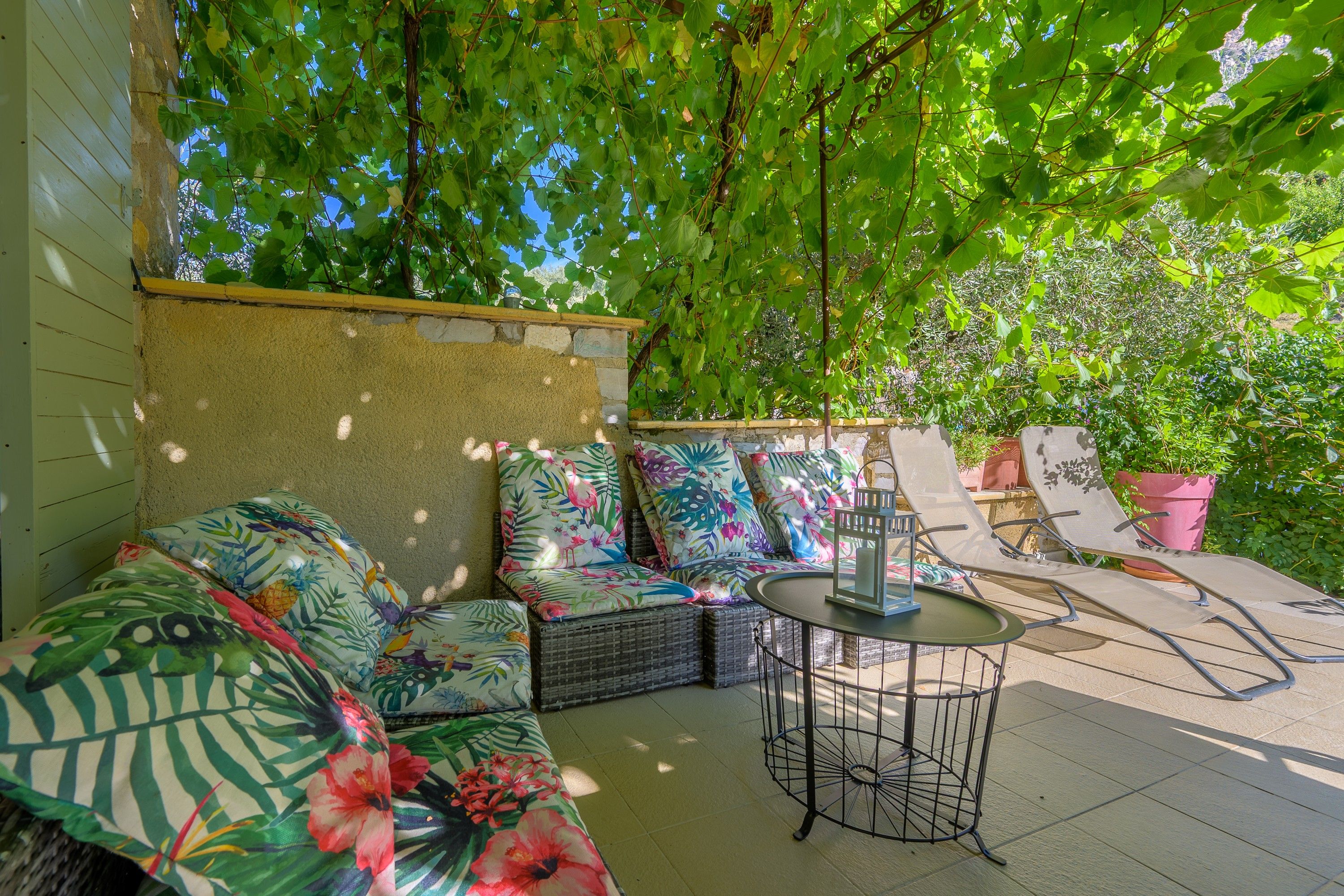 Een overdekt terras met een rieten bank en stoelen, een kleine tafel, en een pergola erboven.