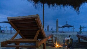 A wooden lounge chair on a sandy beach at dusk with a thatched umbrella overhead and a lantern nearby. - Villa Dyrrahu (Golem)