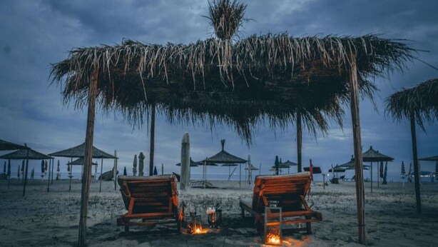 A beach with two wooden chairs under a thatched roof, a fire between them, and several beach umbrellas in the background. - Villa Dyrrahu (Golem)