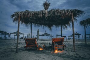 A beach with two wooden chairs under a thatched roof, a fire between them, and several beach umbrellas in the background. - Villa Dyrrahu (Golem)