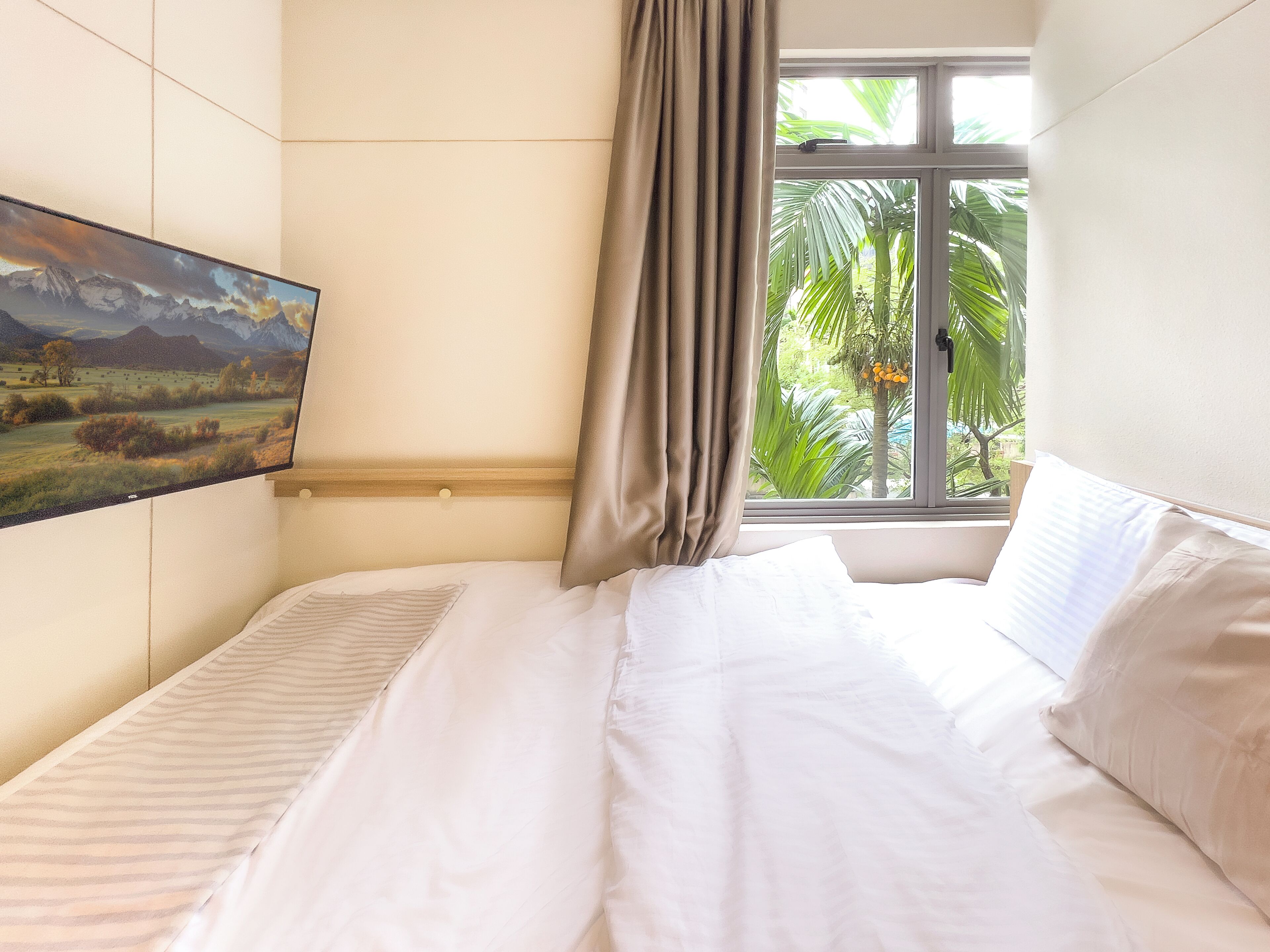 A neatly made bed with white linens in a room with a large window, a painting on the wall, and a curtain partially drawn.