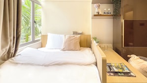 A hotel room with a bed, a bedside table with a magazine, a small shelf with toiletries, and a window with curtains. - Coliwoo Orchard (Singapore)