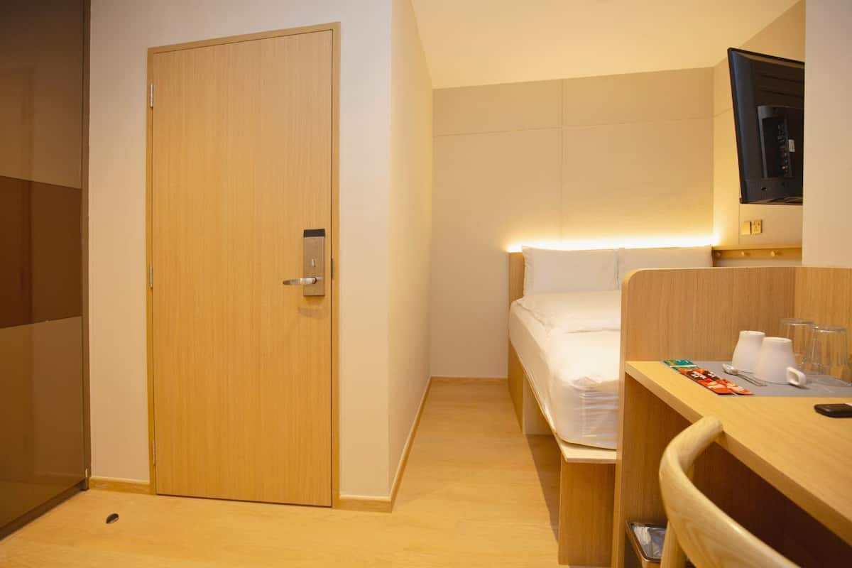 A hotel room with a wooden door, a bed with white bedding, a bedside table with a cup and glasses, and a small trash bin.
