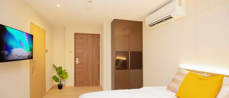 A hotel room with a bed, a television mounted on the wall, a potted plant, a wardrobe, and an air conditioning unit.