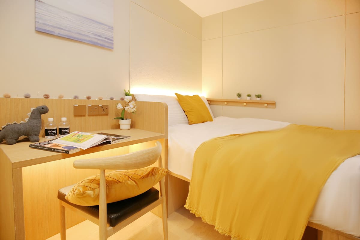 A hotel room with a bed, a desk with a chair, a small shelf with decorative items, and a wall-mounted artwork.