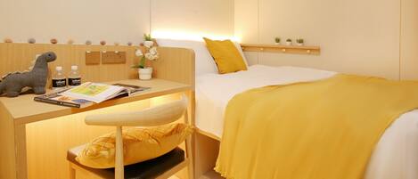 A hotel room with a bed, a desk with a chair, a small shelf with decorative items, and a wall-mounted artwork.
