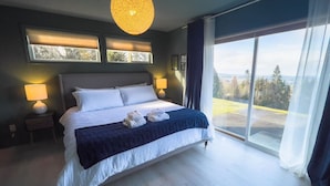 Ein Schlafzimmer mit einem großen Bett, zwei Nachttischlampen, einer Deckenleuchte und Blick durch ein großes Fenster.
