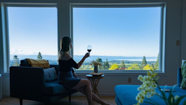 Ein Zimmer mit einem großen Fenster, einem Sofa, einem kleinen Tisch mit einer Vase und Blick auf eine Landschaft.