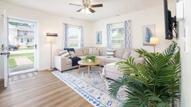 A spacious living room with a beige sofa, a coffee table, and a ceiling fan. There are two windows with curtains, a door leading outside, and a wall-mounted TV.
