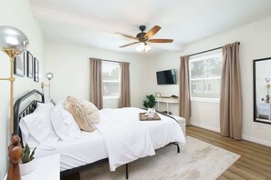 Une chambre avec un lit, un bureau, un ventilateur de plafond et un téléviseur fixé au mur.