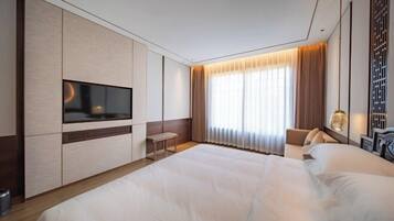 A modern hotel room with a large bed, a flat-screen TV mounted on the wall, a small seating area with a bench, and a large window with curtains.