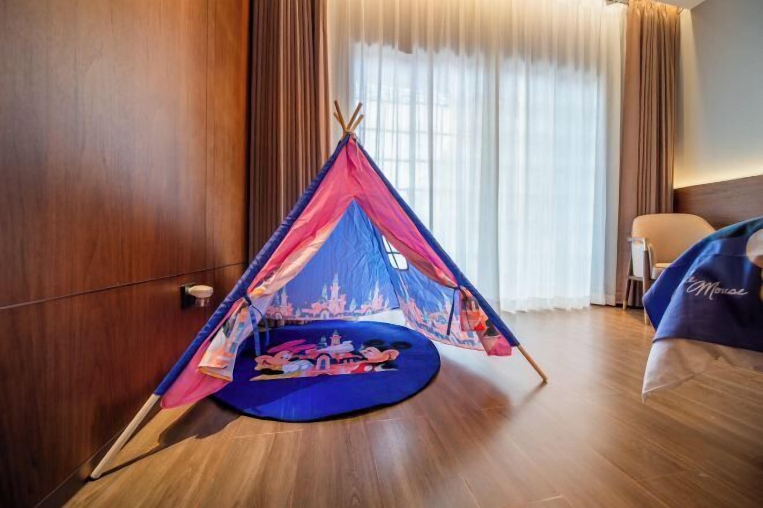 A room with a large window, a bed with a blue blanket, and a tent-like structure with Mickey Mouse on the floor.