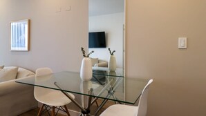 A modern living room with a glass dining table, white chairs, a sofa, and a flat-screen TV mounted on the wall.