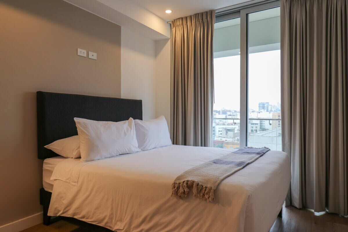 Habitación de hotel con una cama grande, cabecera oscura y vistas a un paisaje urbano a través de amplias ventanas con cortinas.