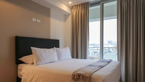 A hotel room with a large bed, a dark headboard, and a view of a cityscape through large windows with curtains.