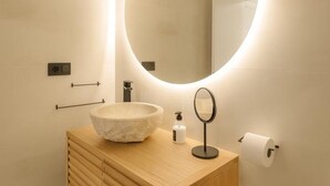 A modern bathroom with a round mirror, a wooden vanity, a white basin, a soap dispenser, and a toilet paper holder.