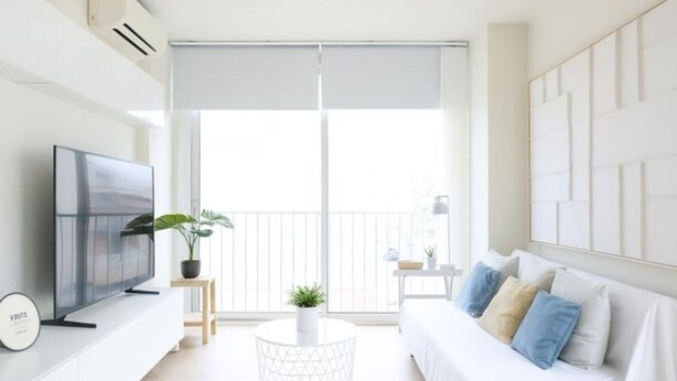 A modern living room with a flat-screen TV, a round coffee table with a plant, a white sofa with cushions, and a large window with a view of the outside.