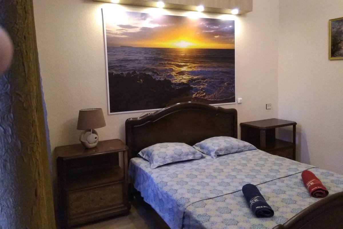 A bedroom with a bed, a nightstand, a lamp, and a wall-mounted picture of a sunset over the ocean.