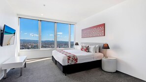 A modern hotel room with a large bed, a desk, and a view of a cityscape through large windows.