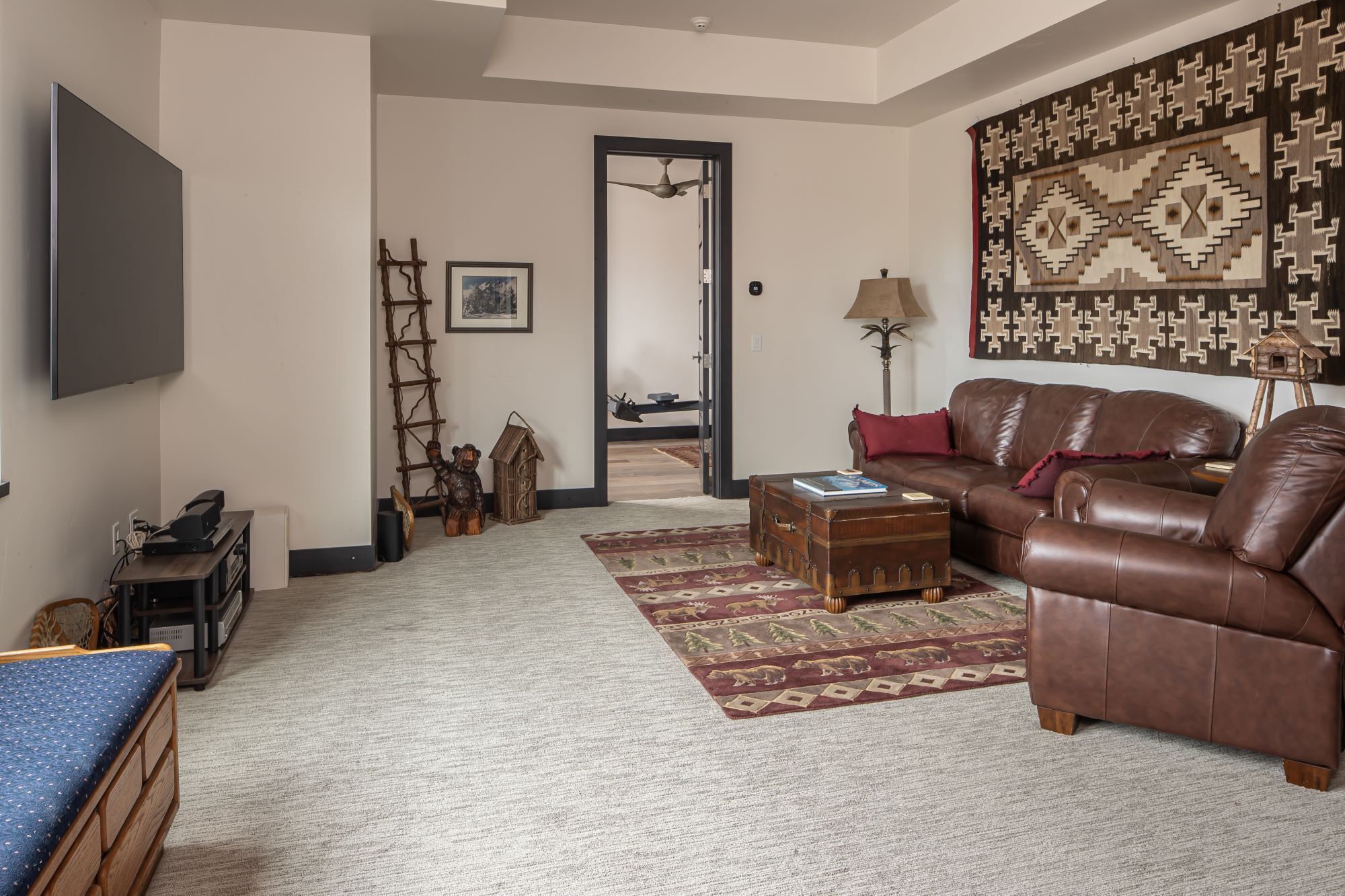 Un salon avec un canapé en cuir marron, un téléviseur à écran plat, une échelle en bois, une statue d’ours, une malle en bois et un tapis à motifs.