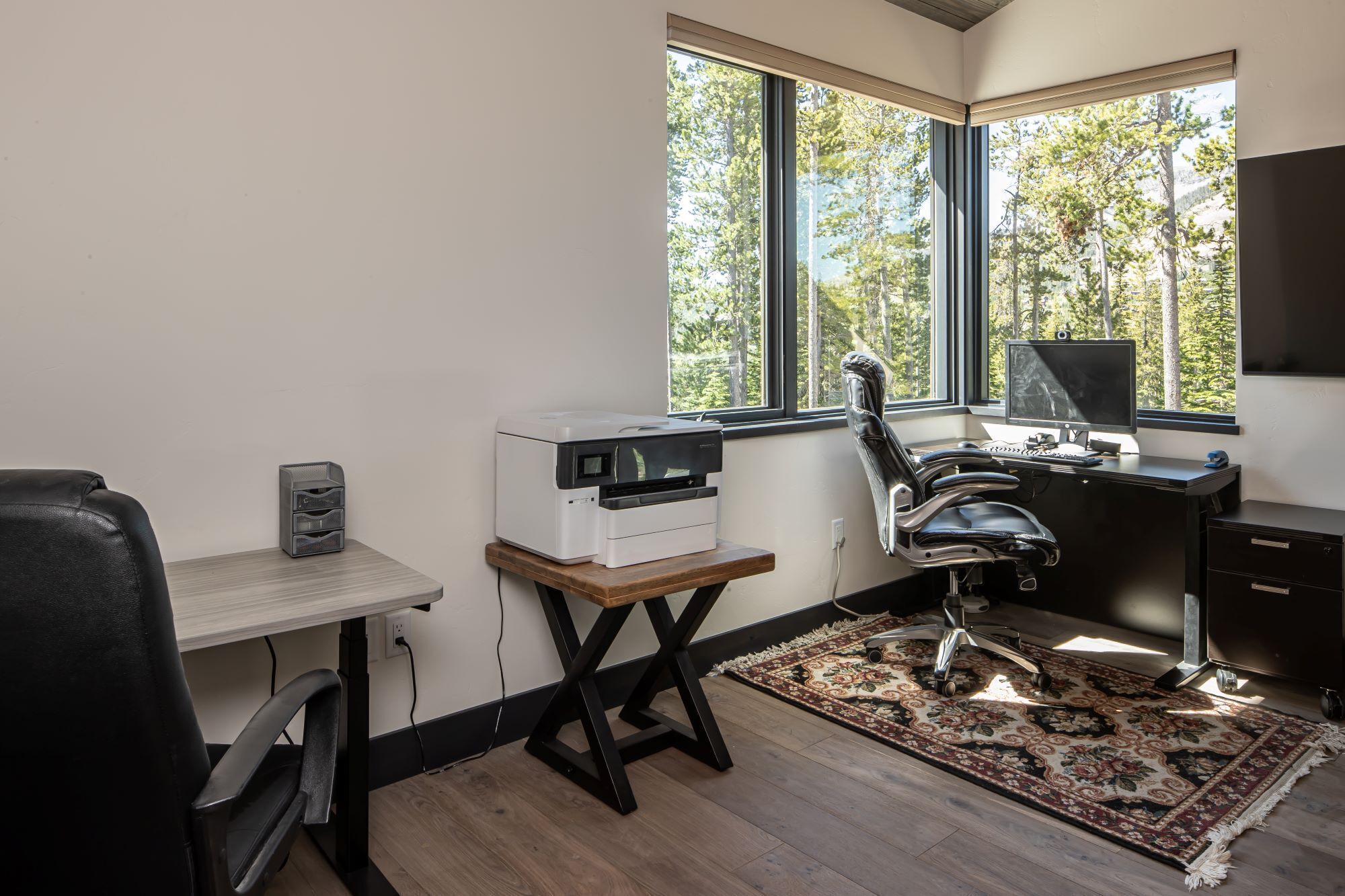 Oficina moderna con impresora, computadora y silla ergonómica. Cuenta con ventanas amplias que dan a vista de árboles y piso de madera.