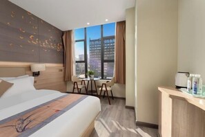Quarto de hotel com uma cama grande, uma mesinha com cadeiras, uma janela vista para a cidade e uma escrivaninha com produtos de higiene pessoal.