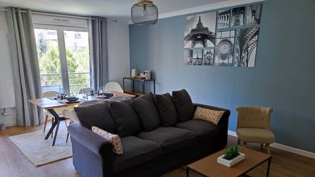 A living room with a dark-colored sofa, a small table with a plant, a chair, and a dining area with a table set for two.