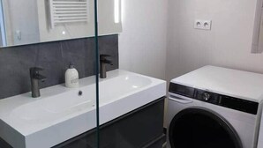 A modern bathroom with a glass partition, a white sink, a mirror, a black faucet, a white bottle, and a white washing machine.