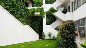 Un edificio moderno con paredes blancas y hiedra verde, un área con césped y pelotas dispersas, y una terraza de madera.