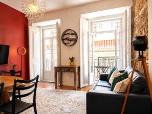 Una sala de estar con un sofá, una mesa de comedor con sillas, un televisor, un reloj de pared y una ventana con vistas.