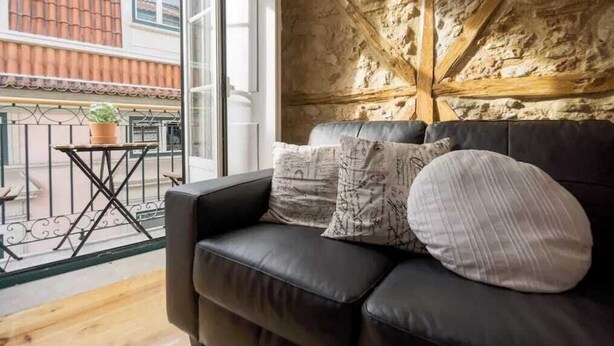 Ein gemütliches Wohnzimmer mit einer schwarzen Ledersofa, gemusterten Kissen und Blick auf eine Steinmauer und einen Balkon.