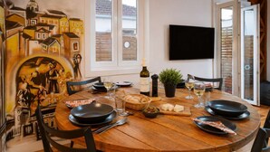 Een eethoek met een ronde houten tafel, zwarte borden, wijnglazen en een fles wijn. Er is een televisie aan de muur bevestigd en een raam met jaloezieën.