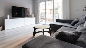 A modern living room with a flat-screen TV on a white cabinet, a grey sofa, a small round table, and a balcony with a metal railing. - Grand House in the Heart of Puerto - Modern, Renovated, Near Beach, 3 Terraces, WIFI (Puerto de Sagunto)