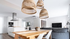 A modern kitchen with white cabinets, a wooden dining table, and a large TV in the living area. - Grand House in the Heart of Puerto - Modern, Renovated, Near Beach, 3 Terraces, WIFI (Puerto de Sagunto)