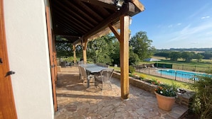Un porche cubierto con piso de piedra, una mesa y sillas, y vistas a una piscina y paisaje verde.